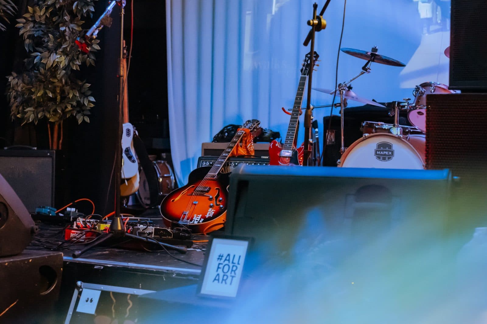 Stage setup with guitars, drums, and an All For Art sign in the foreground
